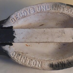 May include: A vintage duck decoy, painted in black, white, and gray. The underside features the embossed text "DECOYS UNLIMITED" and "CLINTON". A wooden support structure is attached to the decoy's base.