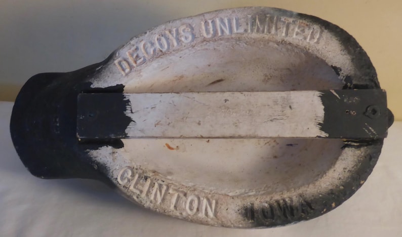 May include: A vintage duck decoy, painted in black, white, and gray. The underside features the embossed text "DECOYS UNLIMITED" and "CLINTON". A wooden support structure is attached to the decoy's base.