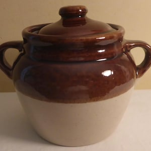 May include: A two-toned ceramic jar with a lid. The top half is a glossy brown, and the bottom half is a cream color. It has two handles and a knob on the lid. The jar appears to be a vintage kitchen item.