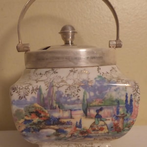 May include: A vintage ceramic biscuit jar with a silver handle and lid. The jar features a hand-painted landscape scene in pastel colors, including a bridge, trees, and figures. The jar rests on three small feet.