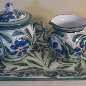 Peut inclure: Un sucrier en céramique avec couvercle, une crémière et un plateau de service. L'ensemble est décoré de motifs floraux bleus et de feuilles vertes sur fond blanc. Le plateau est hexagonal avec une bordure verte.
