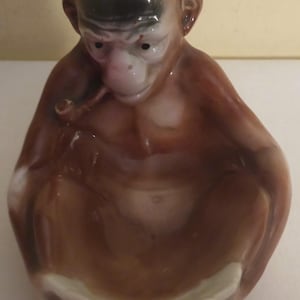 May include: A brown ceramic monkey figurine with a pipe in its mouth, sitting with its legs crossed and holding a small dish in its lap. The monkey has a black face and a white belly.