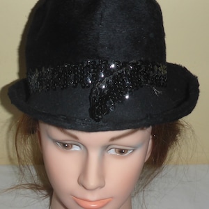 May include: A black cloche hat with a rounded crown and a small brim. The hat features a band of black sequins around the crown, adding a touch of sparkle. The hat is displayed on a mannequin head.