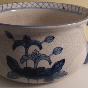 May include: A white ceramic chamber pot with a blue floral design and a blue handle. The pot has a crackled glaze.