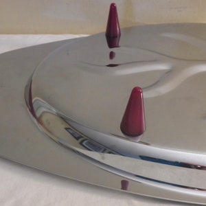 May include: A silver serving tray with a raised, curved design in the centre. The tray has three burgundy coloured, cone-shaped feet.