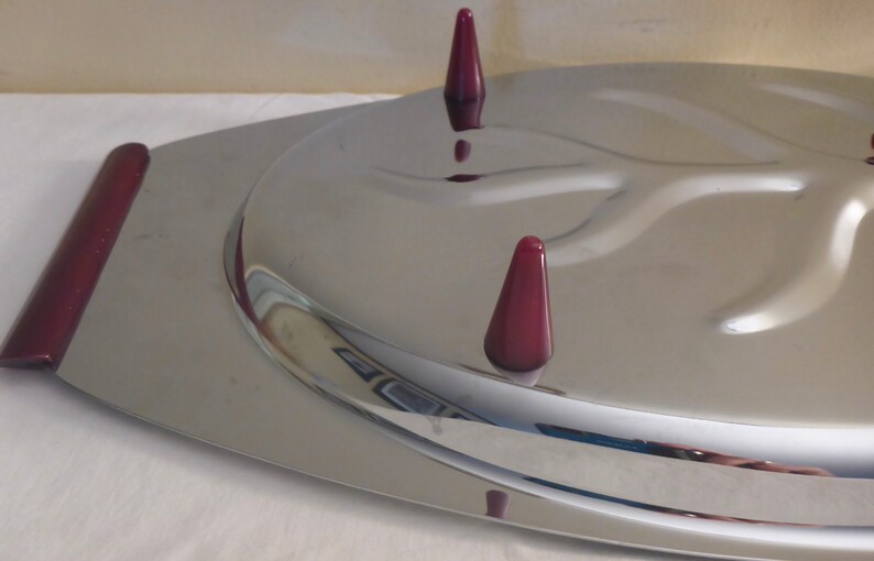 May include: A silver serving tray with a raised, curved design in the centre. The tray has three burgundy coloured, cone-shaped feet.