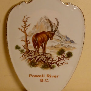 May include: A white ceramic plaque with a brown mountain scene and a brown mountain goat. The plaque has a gold trim and the text "Powell River B.C." in brown.