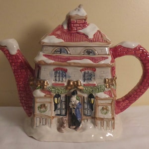 May include: A ceramic teapot shaped like a snow-covered house with a red brick chimney and a green wreath on the door. The teapot has a red brick spout and handle and features two figures standing in front of the house.