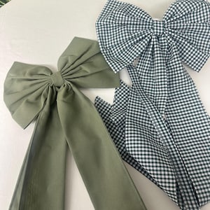 Puede incluir: Dos lazos de tela para el cabello. Uno es verde oliva, el otro tiene un estampado de cuadros blanco y negro. Ambos lazos tienen colas largas y fluidas. Los lazos probablemente sean para peinados.