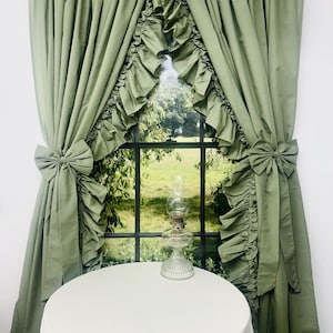 Puede incluir: Cortinas de ventana verde salvia con bordes con volantes y lazos. Las cortinas enmarcan una ventana con vista a un paisaje verde. Un mantel blanco cubre una mesa redonda y una lámpara de cristal está encima.