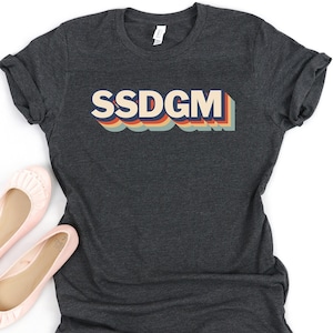 Peut inclure: Un t-shirt gris foncé avec le texte "SSDGMT" dans une police arc-en-ciel rétro.