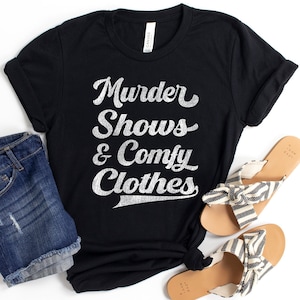 Könnte beinhalten: Ein schwarzes Kurzarm-T-Shirt mit dem weißen, abgenutzten Schriftzug "Murder Shows & Comfy Clothes" auf der Vorderseite. Das T-Shirt ist mit blauen Jeansshorts und einem Paar beigefarbener Sandalen mit schwarz-weiß gestreiften Stoffriemen kombiniert.
