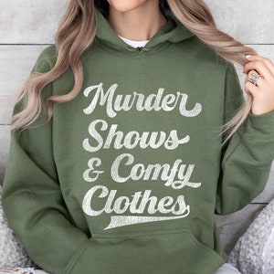 May include: An olive green hoodie featuring the text "Murder Shows & Comfy Clothes" in white, cursive lettering. The hooded sweatshirt has a drawstring and a front pocket. The material appears soft and comfortable.