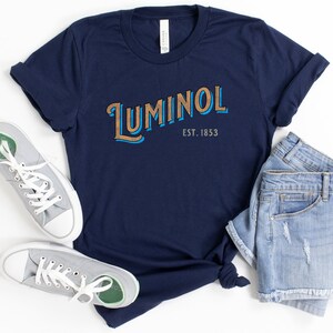 May include: A navy blue t-shirt with the text "Luminol Est. 1853" in a vintage style font. The text is in brown and blue.