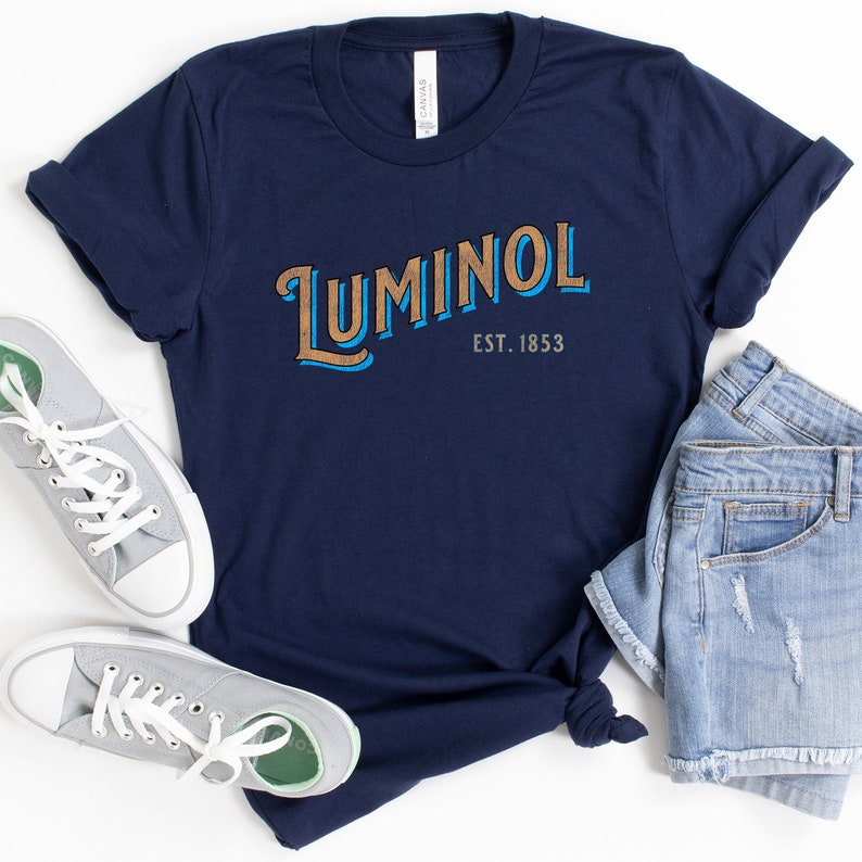 May include: A navy blue t-shirt with the text "Luminol Est. 1853" in a vintage style font. The text is in brown and blue.