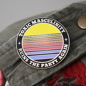 May include: A black and white enamel pin with a yellow sun and colorful stripes. The text around the sun reads "Toxic Masculinity Ruins The Party Again".