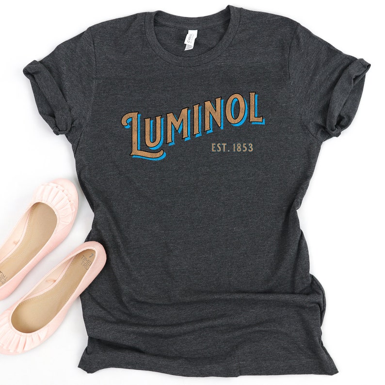 May include: A dark gray t-shirt with a vintage-style logo that reads "Luminol Est. 1853" in blue and brown lettering.