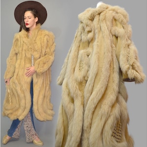 Puede incluir: Un abrigo largo de piel de color marrón claro con un diseño texturizado. El abrigo se muestra en un maniquí y lo lleva una persona. El abrigo tiene un cuello ancho y una silueta fluida. La persona lleva un sombrero marrón, vaqueros azules y pantalones estampados.