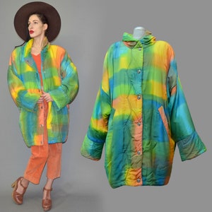 May include: A colorful, oversized coat with a vibrant, abstract design in shades of blue, green, yellow, and orange. The coat features a button-up front, pockets, and a high collar. The model is wearing orange pants and a brown hat.
