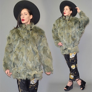 May include: A faux fur coat in shades of green and gold, featuring a high collar and a wide, oversized fit. The coat is paired with black pants with gold celestial designs and a wide-brimmed black hat. The outfit is completed with black platform sandals.