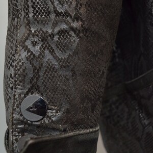 May include: A dark brown jacket sleeve featuring a textured snakeskin pattern throughout the material. The cuff is finished with a single round black button that has a triangular cutout detail, and the fabric appears to have a matte finish.