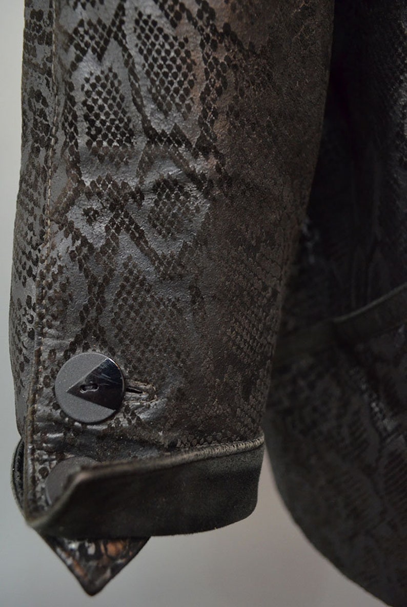 May include: A dark brown jacket sleeve featuring a textured snakeskin pattern throughout the material. The cuff is finished with a single round black button that has a triangular cutout detail, and the fabric appears to have a matte finish.