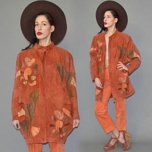 Op de afbeelding: Roestkleurige suède jas en bijpassende broek. De jas heeft bloemen en geometrische patchwork in tinten bruin, oranje en goud. De outfit wordt gecombineerd met een bruine hoed met brede rand en plateauhakken, wat een vintage esthetiek creëert.