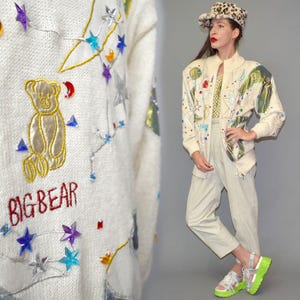Op de afbeelding: Crèmekleurige trui met een gouden beer en de tekst "BIG BEAR" geborduurd in rood. De trui is versierd met kleurrijke ster- en driehoekaccenten. Een persoon draagt de trui met een lichtgekleurde broek, een patroonhoed en neon groene sandalen.