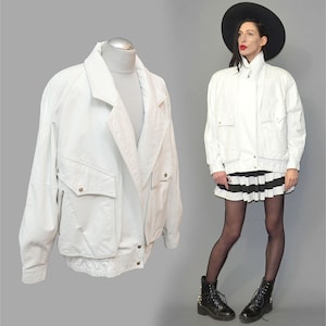 May include: A white leather bomber jacket featuring a wide collar, long sleeves, and two large front pockets with buttoned flaps. The jacket has a ribbed hem and cuffs, a front zipper closure, and metallic hardware accents on the pockets and hem.