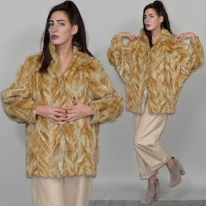 May include: A faux fur coat in shades of tan and cream, featuring a textured, leaf-like pattern. The coat has a wide collar and long sleeves. The model is wearing beige pants and boots.