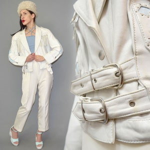 May include: A white leather jacket featuring long fringe detailing on the chest and shoulders, silver-toned metal studs, and two adjustable buckled straps at the waist. The jacket is paired with matching white trousers and light blue platform sandals.