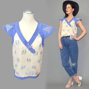 May include: A cream-colored sleeveless top featuring a blue V-neck collar, blue cap sleeves with triangular accents, and a repeating geometric square pattern in blue and gold. The garment includes a decorative hanging tassel detail at the neckline.
