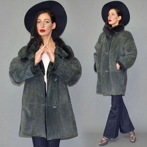 Op de afbeelding: Een lange, donkergroene suède jas met een bontkraag en manchetten. De jas heeft een knoopsluiting aan de voorkant en zakken. Een zwarte hoed met brede rand wordt bij de jas gedragen. Het model draagt een donkerblauwe flared jeans.