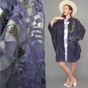 Puede incluir: Una chaqueta azul oscuro con un diseño de patchwork de paneles de ante y cuero texturizado, acentuado con intrincados bordados florales. La prenda tiene cierre frontal de botones, mangas anchas y una silueta holgada y drapeada hasta el muslo.