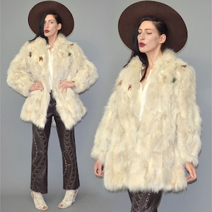 May include: Cream faux fur coat with a wide collar and long sleeves. The coat features decorative accents and is worn with brown leather trousers and a wide-brimmed brown hat. The outfit is completed with open-toe heels.