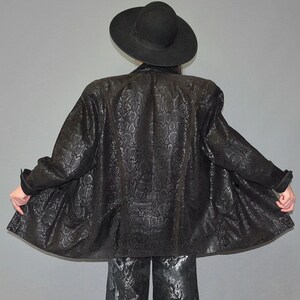 May include: A black blazer featuring a tonal snakeskin pattern with structured shoulders and long sleeves, paired with a wide-brimmed black hat and matching black trousers with a subtle metallic sheen, captured from a rear-facing perspective.