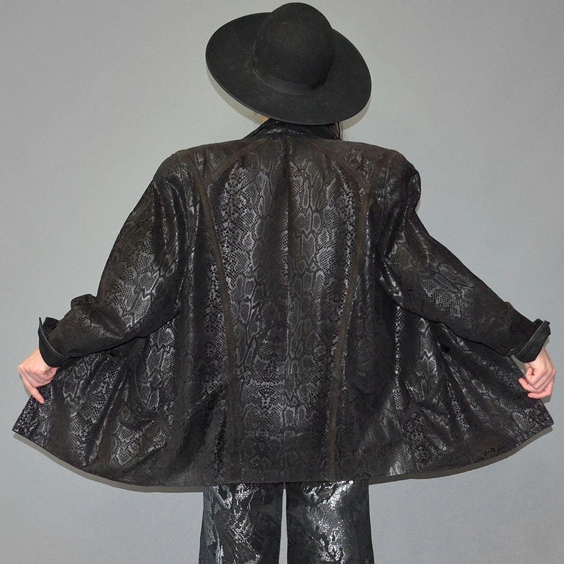 May include: A black blazer featuring a tonal snakeskin pattern with structured shoulders and long sleeves, paired with a wide-brimmed black hat and matching black trousers with a subtle metallic sheen, captured from a rear-facing perspective.