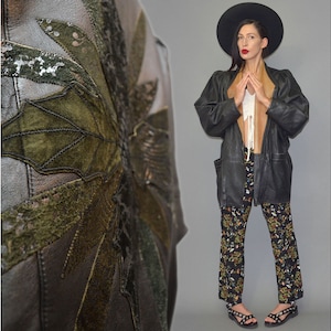 May include: A black leather jacket featuring a large green leaf appliqué with sequin detailing on the back, paired with a tan inner collar, a white top, black patterned trousers, a wide-brimmed black hat, and black strappy sandals.
