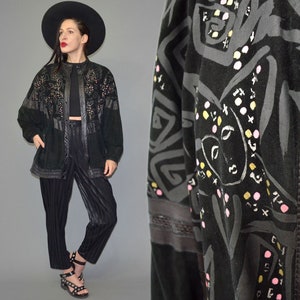 May include: A black jacket featuring a graphic print of abstract faces and geometric shapes in shades of gray, yellow, and pink. The jacket has a button-front closure, long sleeves, and a textured finish. Paired with black pinstriped trousers.