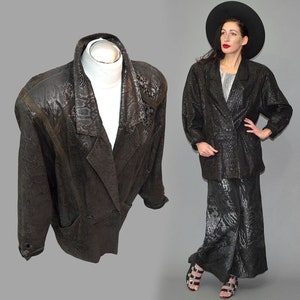 May include: A black blazer featuring a subtle snakeskin texture pattern, wide lapels, and long sleeves. The jacket has a structured shoulder design and a button-front closure. The fabric has a dark, monochromatic finish with a repeating reptile scale motif.