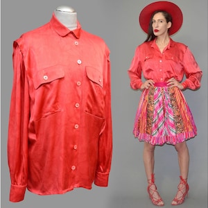 Puede incluir: Una camisa roja brillante con botones y dos bolsillos en el pecho, exhibida en un maniquí. La camisa tiene mangas largas y un cuello clásico. Junto a ella, una persona lleva una camisa roja a juego, una falda estampada y un sombrero rojo.
