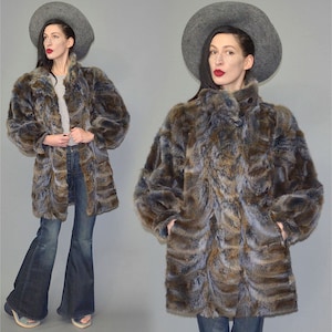 Op de afbeelding: Een lange imitatiebontjas in bruin-, blauw- en grijstinten. De jas heeft een hoge kraag en een knoopsluiting. Het model draagt een grijze hoed en een blauwe jeans. De jas is ongeveer 102 cm lang.