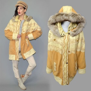 Puede incluir: Un abrigo de invierno color beige y crema con capucha forrada de piel. El abrigo presenta costuras decorativas, cierres de botones y detalles bordados. La persona lleva el abrigo con un sombrero a juego y pantalones blancos.