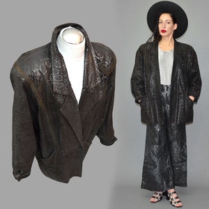 May include: A black blazer featuring a subtle snakeskin texture pattern throughout the fabric, a wide notched lapel, long sleeves, and two front pockets. The jacket has a structured shoulder design and a button closure at the front waist area.