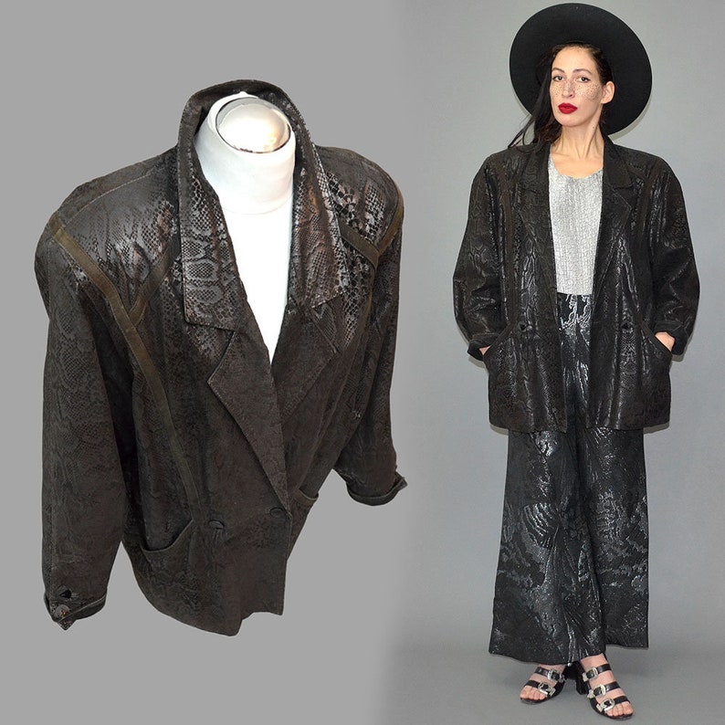 May include: A black blazer featuring a subtle snakeskin texture pattern throughout the fabric, a wide notched lapel, long sleeves, and two front pockets. The jacket has a structured shoulder design and a button closure at the front waist area.