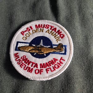 May include: A circular embroidered patch featuring a gold P-51 Mustang airplane silhouette centered on a blue star and white bar emblem. The text reads P-51 MUSTANG GOLDEN ANNIV and SANTA MARIA MUSEUM OF FLIGHT in red and gold thread on a white background.
