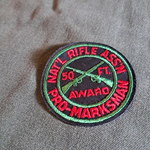 May include: A circular patch with the text "NAT'L RIFLE ASSN" and "PRO-MARKSMAN" in red, with a green border. The center features crossed rifles and the text "50 FT. AWARD". The patch is on a textured, olive-green background.