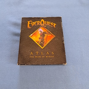 May include: A black rectangular box with a textured surface featuring the EverQuest logo in orange at the top. A central diamond-shaped cutout displays an illustration of a character. Below the image, the text reads ATLAS THE MAPS OF MYRIST.