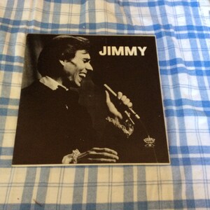Puede incluir: Una foto en blanco y negro de un hombre cantando en un micrófono. La foto está en una portada de álbum de vinilo con la palabra "JIMMY" impresa en letras blancas en la parte superior.