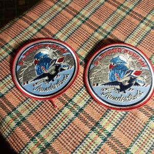 May include: Two round embroidered patches with red, white, and blue accents. The patches feature a blue bird with a red, white, and blue jet plane. The text on the patches reads "A Legend Reborn Thunderbirds".
